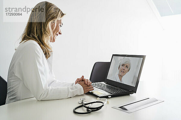 Lächelnde Ärztin im Gespräch mit einem älteren Patienten während einer telemedizinischen Behandlung über einen Laptop am Schreibtisch
