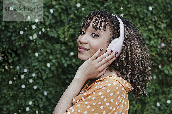 Junge Frau berührt Kopfhörer beim Hören von Musik durch Hecke