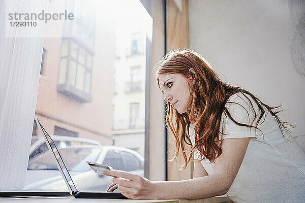 Rothaarige Frau bei der Online-Zahlung per Kreditkarte  während sie einen Laptop vor einem Fenster zu Hause benutzt