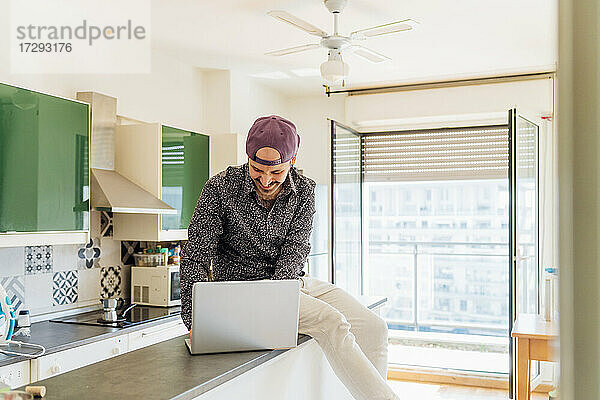Lächelnder junger Mann  der einen Laptop benutzt  während er auf einer Kücheninsel zu Hause sitzt