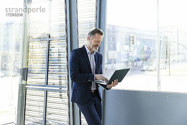 Lächelnder Geschäftsmann arbeitet am Laptop vor dem Fenster im Büro