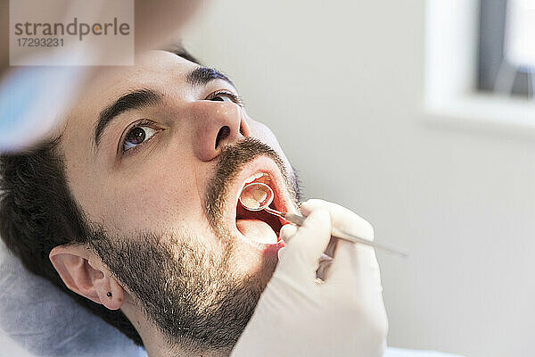 Zahnärztin mit angewinkeltem Spiegel bei der Untersuchung der Zähne eines männlichen Patienten in einer medizinischen Klinik