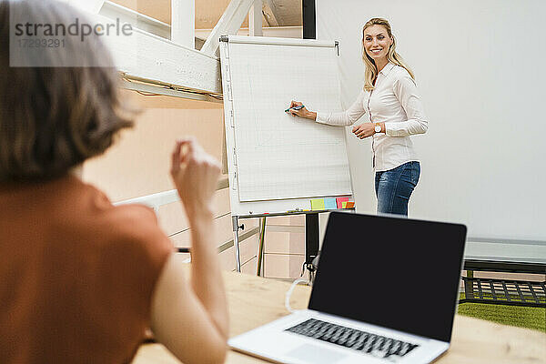 Lächelnde Geschäftsfrau  die einen jungen Kollegen anschaut  während sie auf eine weiße Tafel schreibt und eine Strategie in einem kreativen Büro plant