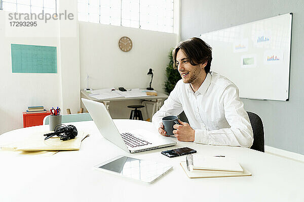 Männlicher Unternehmer mit Kaffeetasse und Laptop bei der Arbeit im Kreativbüro