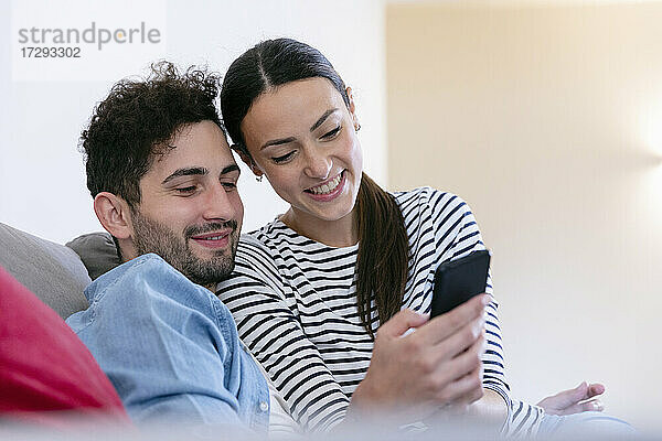 Lächelndes Paar  das im heimischen Wohnzimmer ein Mobiltelefon benutzt