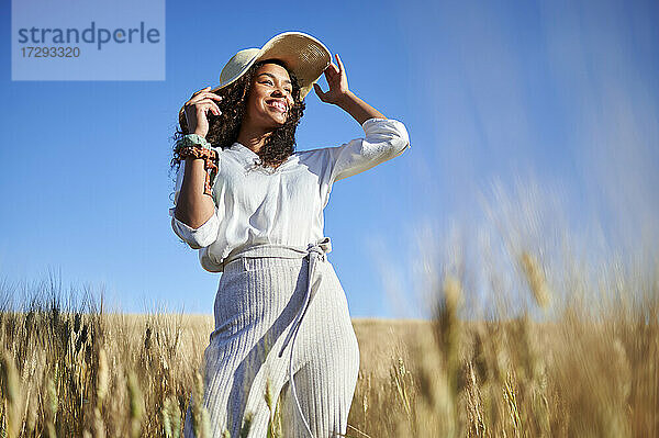 Lächelnde junge Frau mit Hut  die wegschaut  während sie in einem Weizenfeld steht