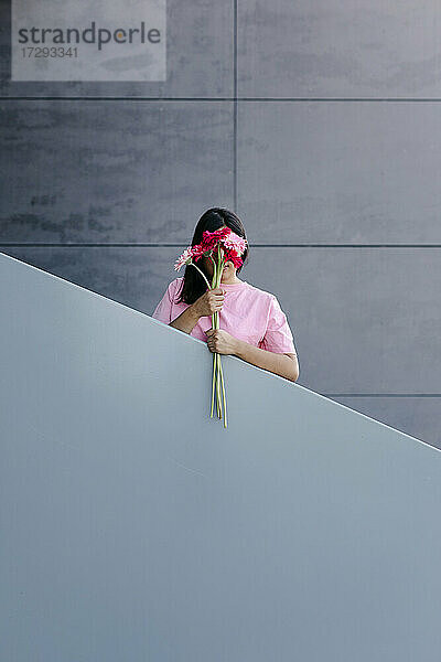 Junge Frau versteckt ihr Gesicht mit Gerbera-Gänseblümchen vor einem Geländer