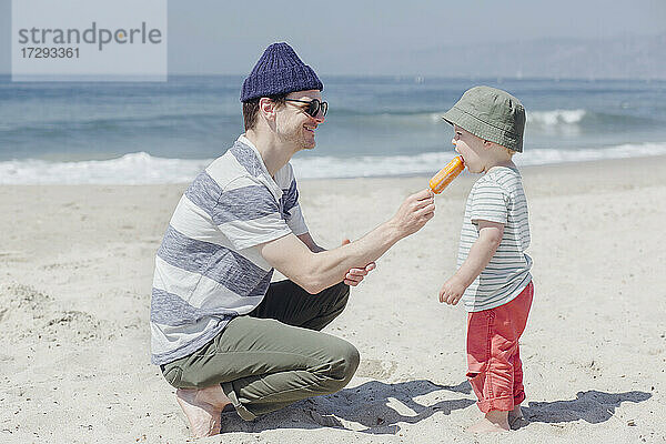 Lächelnder Mann  der seinen Sohn am Strand mit Eiscreme füttert  an einem sonnigen Tag