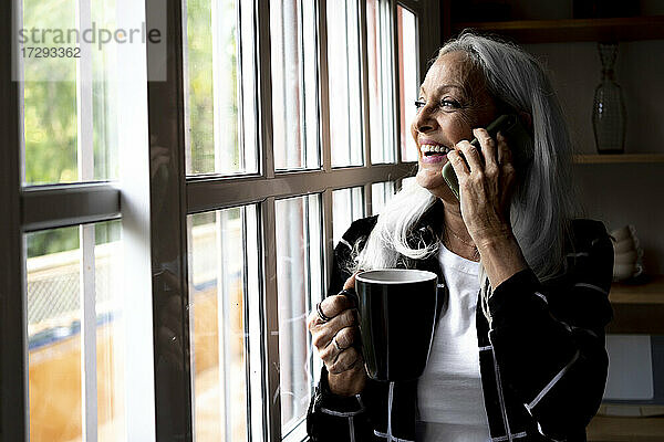 Glückliche Frau mit Kaffeetasse  die durch das Fenster schaut  während sie zu Hause mit ihrem Smartphone telefoniert
