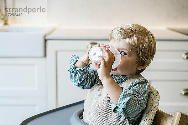 Blonder Junge trinkt Milch  während er auf einem Hochstuhl in der Küche zu Hause sitzt
