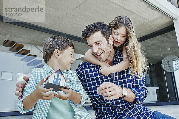 Lächelnder Junge  der ein Hausmodell hält  sitzt neben einem verspielten Vater und einem Mädchen im Hinterhof