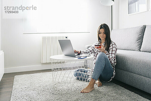 Junge Frau mit Kreditkarte beim Online-Shopping mit Laptop im Wohnzimmer