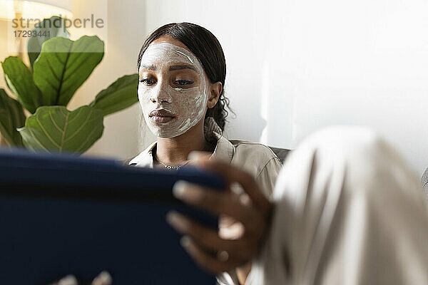 Frau  die ein digitales Tablet benutzt  während sie mit einer Schönheitsmaske im Gesicht zu Hause sitzt