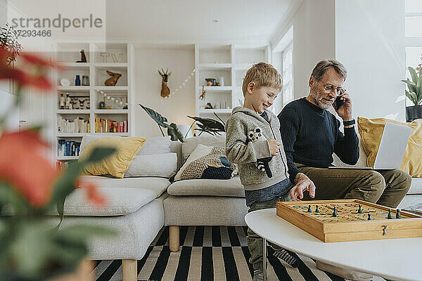 Mann spricht mit seinem Handy  während er mit seinem Sohn zu Hause ein Ludo-Spiel spielt
