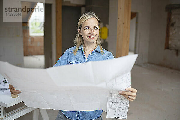 Lächelnde Architektin  die den Blick abwendet  während sie einen Bauplan auf der Baustelle hält