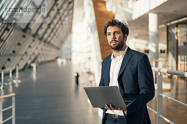 Männlicher Unternehmer mit Laptop  der wegschaut  während er in der Büro-Lobby steht