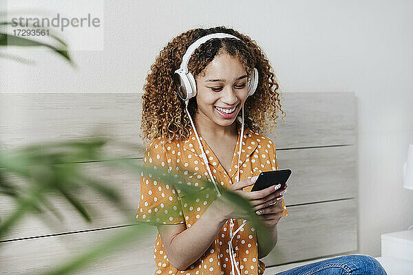 Junge Frau  die ein Mobiltelefon benutzt  während sie auf dem Bett zu Hause Musik hört