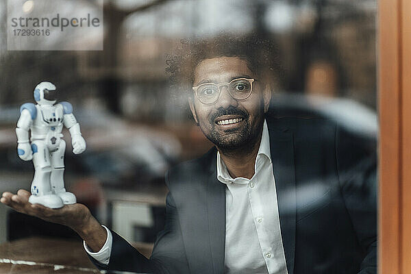 Lächelnder reifer Geschäftsmann  der mit einem Robotermodell in einem Café sitzt  gesehen durch Glas