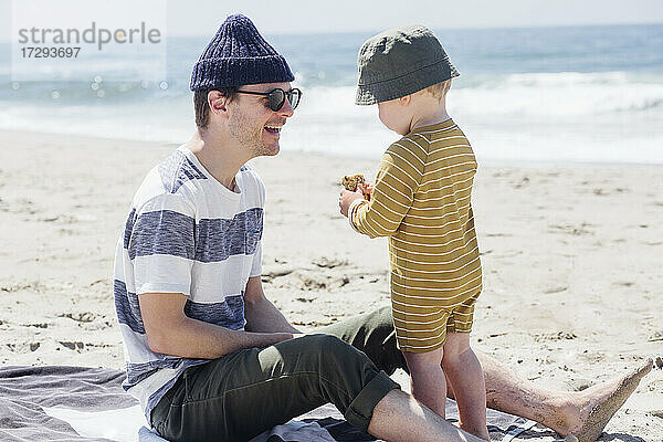 Lächelnder Vater sieht seinen Sohn beim Essen am Strand an