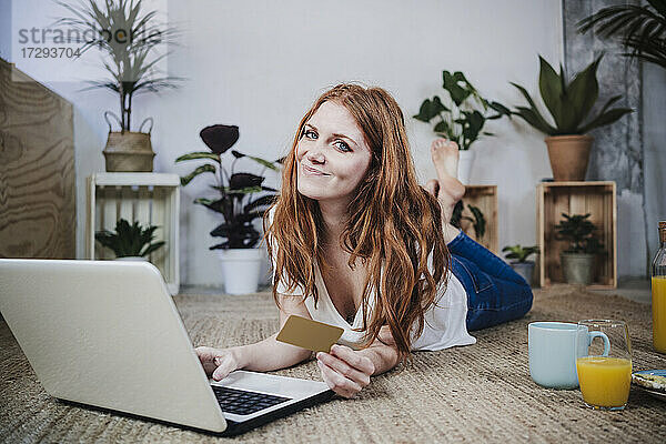 Lächelnde rothaarige Frau mit Kreditkarte beim Online-Shopping auf dem Laptop zu Hause