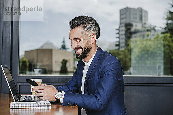 Lächelnder Geschäftsmann mit Laptop und Mobiltelefon in einer Cafeteria