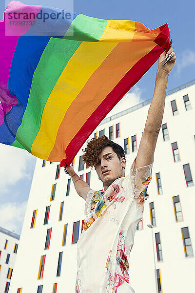 Junger Mann weht Regenbogenflagge vor einem Gebäude