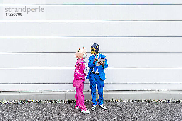 Mann und Frau in farbenfrohen Anzügen und Tiermasken unterhalten sich über ein Klemmbrett vor einer weißen Wand