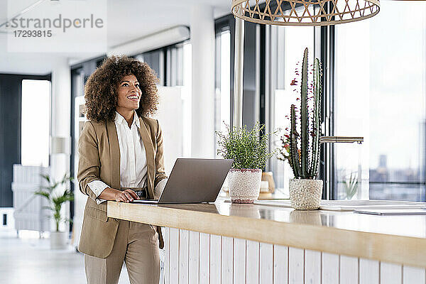 Lächelnde Unternehmerin  die eine Hängelampe betrachtet  während sie in der Cafeteria vor einem Laptop steht
