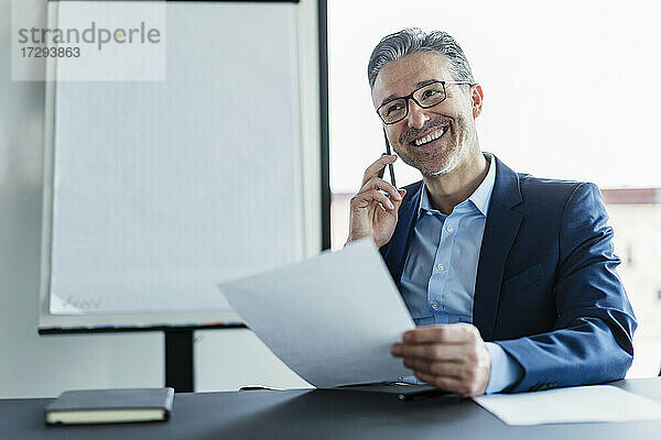 Lächelnder Geschäftsmann mit Brille  der im Büro mit einem Mobiltelefon spricht und wegschaut