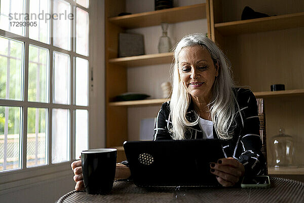 Lächelnde ältere Frau  die auf ein digitales Tablet schaut  während sie zu Hause Kaffee trinkt