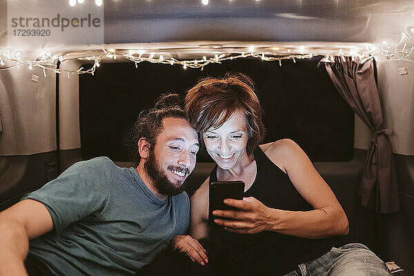 Lächelndes Paar  das in einem beleuchteten Lieferwagen nachts ein Mobiltelefon benutzt