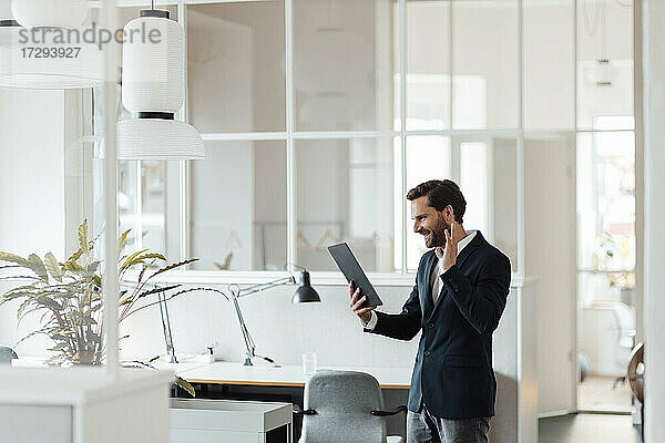 Lächelnder Geschäftsmann winkt mit der Hand zu einem Videoanruf über ein digitales Tablet im Büro