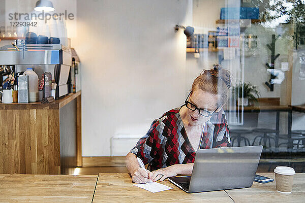 Frau schreibt auf Papier  während sie in einem Café vor einem Laptop sitzt