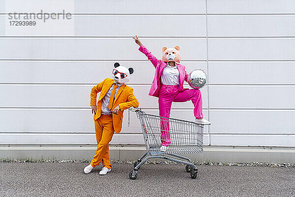 Mann und Frau in farbenfrohen Anzügen und Tiermasken posieren mit Einkaufswagen und Discokugel vor einer weißen Wand
