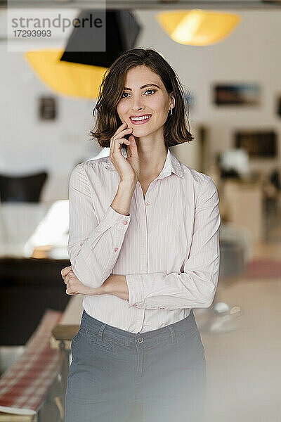 Lächelnde schöne weibliche Fachkraft mit Hand am Kinn im Büro stehend und wegschauend