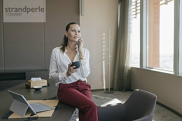 Reife Geschäftsfrau  die eine Kaffeetasse hält  während sie mit einem Smartphone im Büro telefoniert