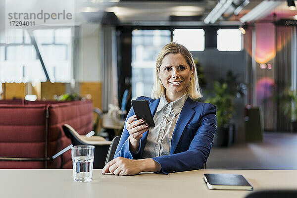 Lächelnde weibliche Fachkraft sitzt mit Handy am Schreibtisch im Büro