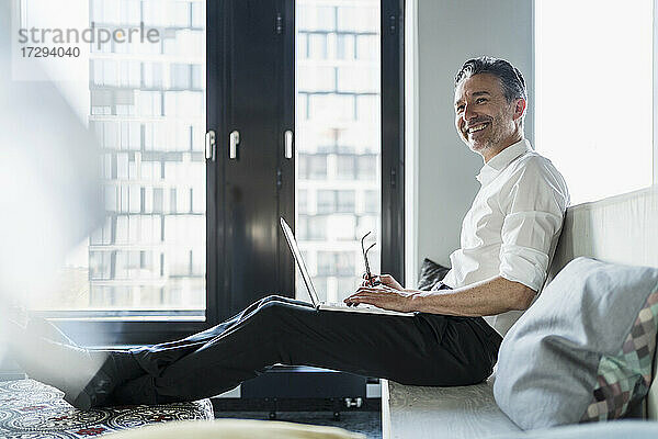 Lächelnder männlicher Unternehmer sitzt mit Laptop auf dem Sitz im Büro und schaut weg