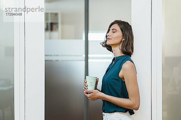 Junge Geschäftsfrau mit geschlossenen Augen  die eine Einweg-Kaffeetasse hält  während sie an der Tür im Büro lehnt