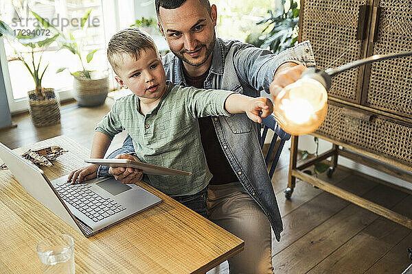 Mann und Sohn zeigen auf Glühbirne  während sie mit Tablet und Laptop im intelligenten Haus sitzen