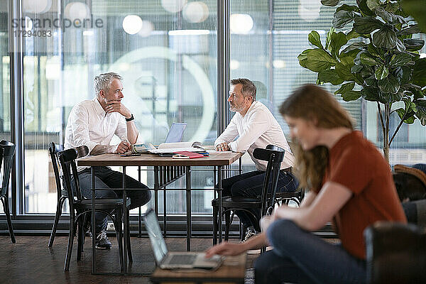 Männliche Unternehmer diskutieren im Hintergrund  während eine Geschäftsfrau im Büro am Laptop arbeitet