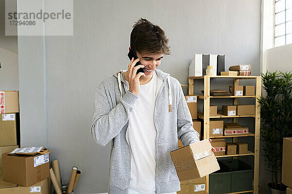 Lächelnder männlicher Unternehmer  der mit einem Mobiltelefon spricht  während er ein Paket in einem Lagerhaus betrachtet