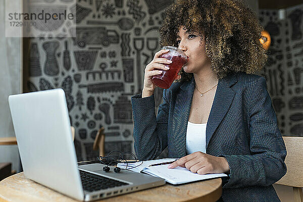 Afro-Geschäftsfrau schaut weg  während sie vor einem Laptop trinkt