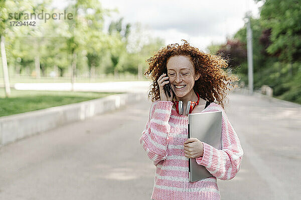Fröhliche junge Frau mit Laptop  die im Park mit ihrem Handy telefoniert