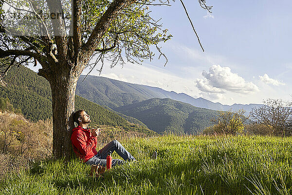 Mann mit roter Jacke lehnt an einem Baum und genießt die Natur