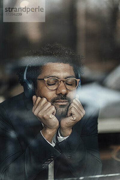 Männlicher Unternehmer mit Händen am Kinn hört Musik über Kopfhörer in einem Café  gesehen durch Glas