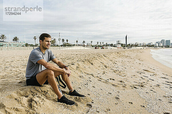 Männlicher Sportler schaut weg  während er am Strand im Sand sitzt