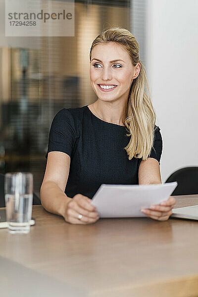 Schöne lächelnde junge Unternehmerin sitzt mit Dokument am Schreibtisch und schaut weg