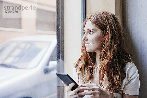 Junge Frau mit Mobiltelefon schaut durch ein Fenster zu Hause