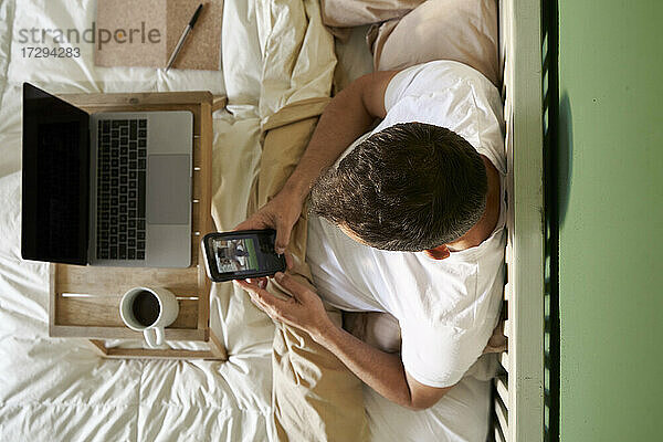 Ein Mann führt einen Videochat mit einem Freund über sein Mobiltelefon  während er im Bett am Laptop sitzt